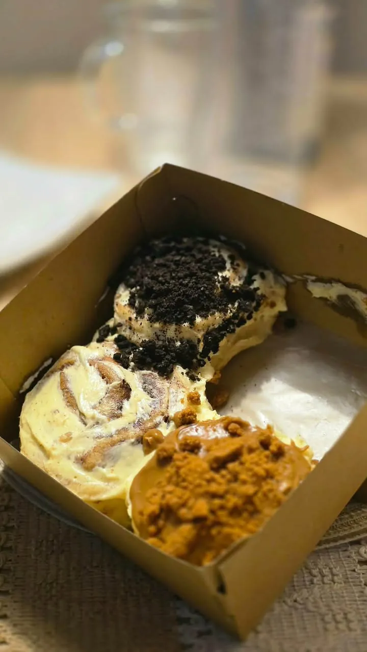 Three homemade cinnamon rolls in a kraft box — one with chocolate cookie crumbs, one with a creamy frosting swirl, one with biscuit crumble