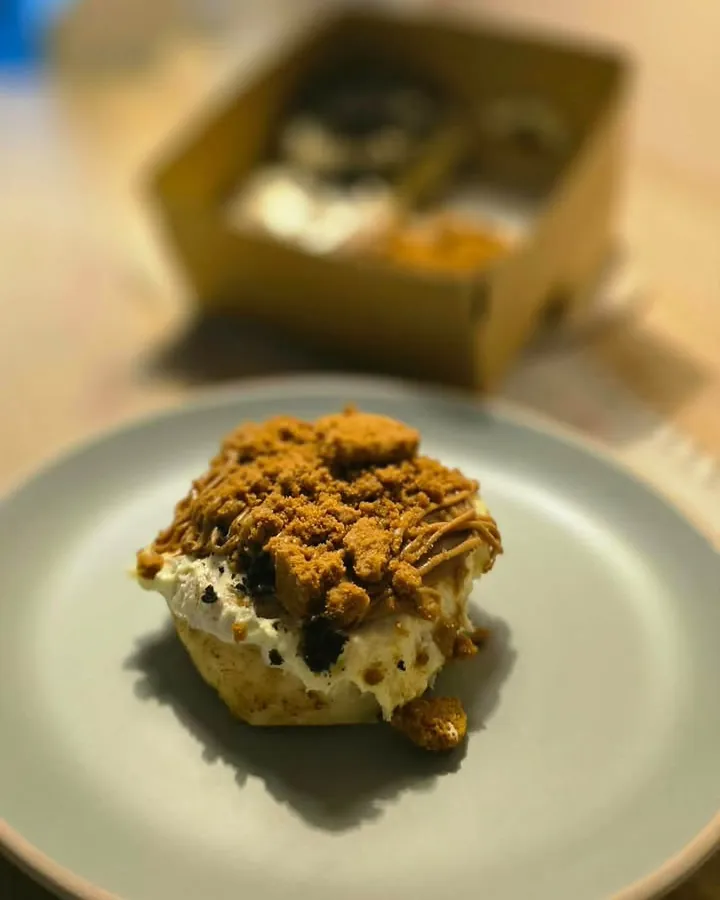 A homemade cinnamon roll topped with biscuit crumble on a plate, with a kraft box of rolls behind it